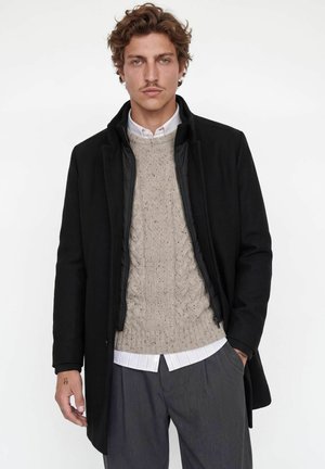 Man with curly hair wearing a black coat, beige cable knit sweater, white shirt, and gray pants standing against a white background.
