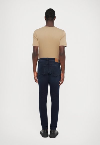 Tricou ajustat de culoare tan, asortat cu blugi skinny de culoare albastru închis. Blugii au o petă de piele maro pe spate, cu un cusătură subtilă și fără modele vizibile.