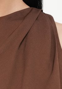 Top marrone asimmetrico realizzato in tessuto morbido e liscio; presenta dettagli drappeggiati sulla spalla e una sottile texture. Nessun sistema di chiusura o decorazione visibile.