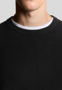 Zwarte gebreide trui met een ronde hals. De stof lijkt glad en zacht, met een contrasterende witte binnenkraag.