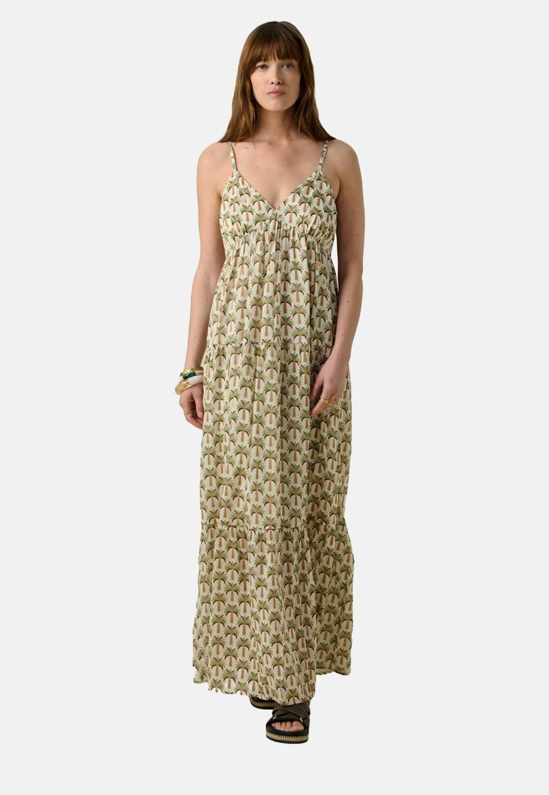 Robe maxi en tissu léger, avec un motif de feuilles vertes et beiges, des bretelles spaghetti et une silhouette à volants. Associée à des sandales sombres.