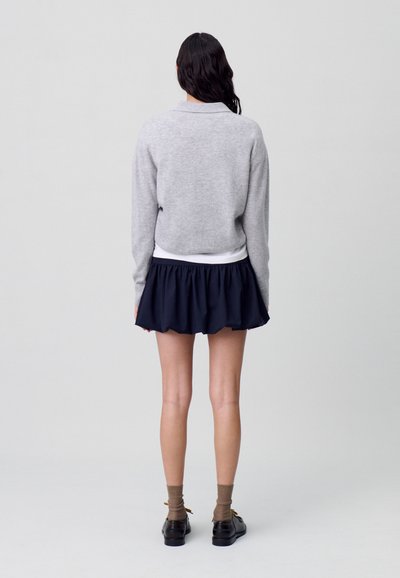 Pull tricot gris à col rond, associé à une jupe plissée marine. La tenue comprend une ceinture blanche et des chaussures noires.