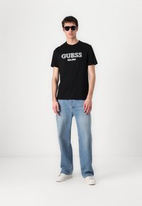 Juoda medvilninė marškinėliai su sidabro spalvos užrašu "GUESS EST. 1981". Sukombinuoti su šviesiai mėlynomis džinsinėmis kelnėmis ir baltomis sportinėmis bateliais, modelis dėvi saulės akinius.