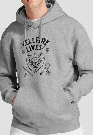 Mann trägt grauen Hoodie mit schwarzem Motiv eines Schädels mit Hörnern als Schild, Schwertern und dem Schriftzug "HELLFIRE LIVES!" auf der Brust. Hände in den Taschen des Hoodies.