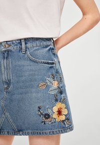 Jeansrok met bloemenborduursel in gele, blauwe en bruine tinten. Heeft een getailleerde taille en zichtbare stiksels, met een knie-lange snit.