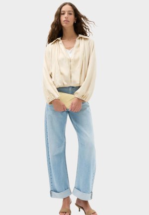 Kvinde med langt krøllet hår iført beige bluse med lynlås, lyseblå opkumpede jeans, brune hælede sandaler og holder en lille beige clutch.
