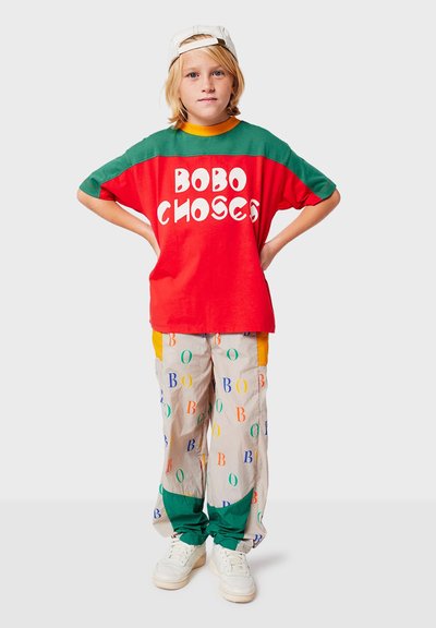 Rød og grøn oversized t-shirt med "BOBO CHOSES" tekst, parret med beige bukser med farverige bogstavmønstre og grønne accenter. Hvide sneakers.