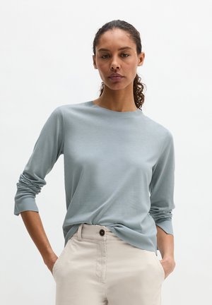 Woman with curly hair wearing a light blue long-sleeve shirt and beige pants, standing with hands in pockets against a plain background.