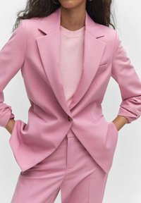 Femme portant un blazer rose clair ajusté et un pantalon assorti, mains dans les poches, avec un haut en maille rose clair en dessous.