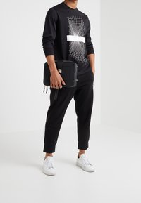 Svart långärmad skjorta med en vit grafisk design, kombinerad med svarta byxor och vita sneakers, hållandes en svart dragkedjeförsedd väska.