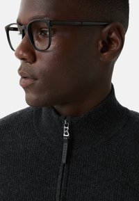 Mężczyzna o ciemnej karnacji, noszący czarne prostokątne okulary i szary sweter z zamkiem błyskawicznym, zwrócony w lewo na białym tle.