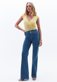 Top de encaje amarillo con mangas cortas, combinado con jeans de campana azules de talle alto que tienen un cierre de botón y detalles cosidos.