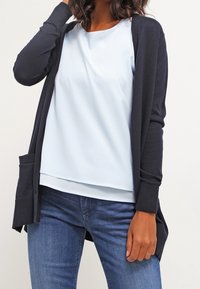 Marinblå cardigan med öppen front och fickor, kombinerad med en ljusblå blus. Blusen har en mjuk tygkvalitet och en avslappnad passform.