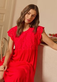 Femme aux longs cheveux bruns portant une robe rouge à œillets, assise sur une chaise et appuyée sur une table dans une pièce doucement éclairée.