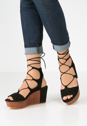 Kobieta ubrana w ciemnoniebieskie, podwinięte dżinsy i czarne zamszowe sandały na koturnie z drewnianą platformą oraz krzyżującymi się paskami w okolicy kostki.