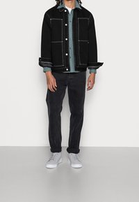 Zwarte spijkerjasje met witte steken, gedragen over een groen shirt. Donkere spijkerbroek en lichtgrijze sneakers maken de outfit compleet.