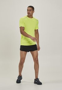 Neon gul atletisk t-shirt med korte ærmer, fugttransporterende stof og reflektive detaljer, kombineret med sorte atletiske shorts og mørke sneakers.