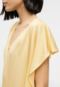 Μπλούζα με μανίκια φτερού σε απαλό κίτρινο ύφασμα με βαθύ V-λαιμόκοψη, που διαθέτει λεπτή draping και ομαλή, ελαφριά υφή.
