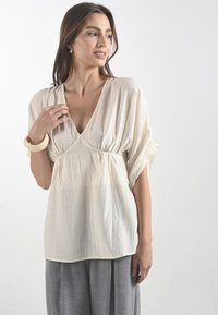 Donna in un top fluente di colore crema con un ampio scollo, abbinato a pantaloni grigi. Indossa un grande bracciale e sorride dolcemente.