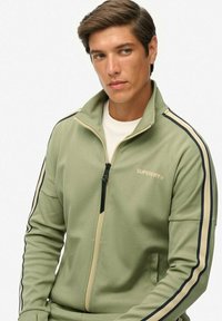 Groene jas met ritssluiting en hoge kraag, met contrasterende strepen langs de mouwen en een merklogo op de borst. Gladde textuur.