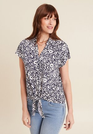 Frau trägt eine marineblaue Bluse mit kurzen Ärmeln und weißem Blumenmuster, die an der Taille gebunden ist, kombiniert mit hellblauen Jeans, stehend vor einem beigen Hintergrund.