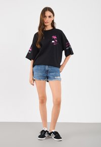 Tricou negru oversized cu grafică de palmier roz, asortat cu pantaloni scurți din denim cu aspect uzat și teniși negri cu dungi albe.