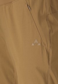 Nahaufnahme von hellbraunen Stoffhosen mit einer diagonalen Reißverschlusstasche und einem kleinen dreieckigen Logo in der Nähe der Taschennaht.