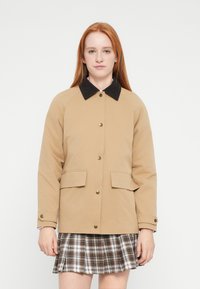 Ανοιχτό καφέ μπουφάν με γιακά από μαύρο κορντόρι, κουμπιά τύπου snap και δύο μπροστινές τσέπες. Φοριέται πάνω από καρό φούστα. Λεπτή υφή.