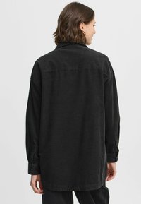 Camisa de corduroy negra con un corte relajado, mangas largas y cuello. Presenta un puño abotonado y un detalle de costura en el yugo trasero.