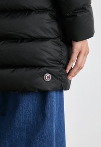 Zwarte pufferjack met een gestikt ontwerp, voorzien van een ronde logo-embleem bij de zoom en een gladde, glanzende textuur. Gepaarde met denim onderkant.