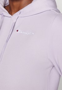 Lavendel Champion hoodie med dragsnören, broderad logotyp på bröstet, bärs av en person med medel-mörk hudton.