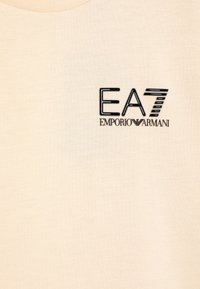 Krämfärgad bomullströja med en svart tryckt logotyp med texten "EA7" och "EMPORIO ARMANI". Slät textur, design med rund hals.