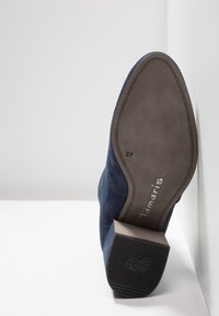 Chaussure en daim bleu marine vue de la semelle, taille 37, avec marquage "Tamaris" et talon antidérapant, appuyée contre une surface d'angle blanche.