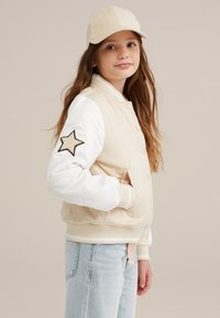 Bežinė bomber tipo striukė su baltomis rankovėmis, pasižyminti juoda žvaigždute ir raštuotais rankogaliais. Derinama su bežine kepure ir šviesiai mėlynomis džinsais.