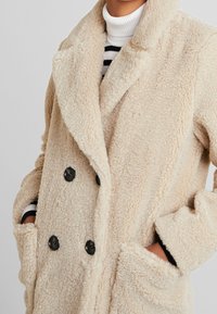 Abrigo beige cruzado con textura suave, grandes botones negros y bolsillos laterales, con cuello chal y corte holgado.