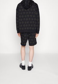 Haut à capuche noir et shorts avec un imprimé de smiley jaune, comprenant une capuche, des poignets côtelés et un tissu texturé ; associés à des chaussures noires.