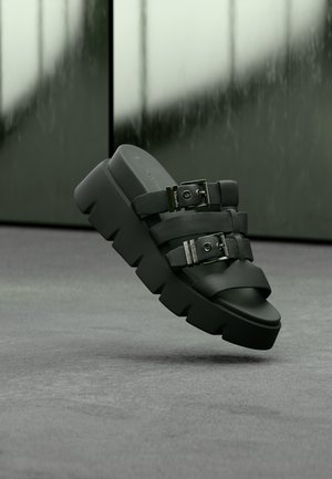 Sort robust slip-sandal med tyk sål og to justerbare stropper med sølvspænder, sat mod en sløret grøn-grå baggrund.