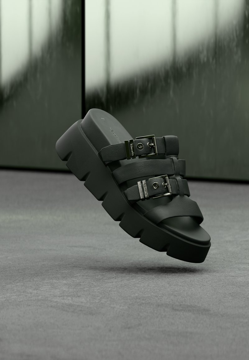 Sandale noire à semelle épaisse, avec deux brides réglables à boucles argentées, sur un fond flou vert-gris.