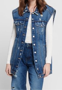 Jeansväst i ljusblå färg, med slitage, två bröstfickor och metallknappar. Lager på en vit långärmad topp.