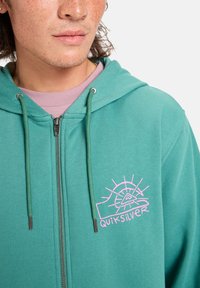 Tyrkysová mikina na zips s ružovým grafickým logom s dizajnom slnka. Obsahuje šnúrky na stiahnutie a kovové detaily zipsu.