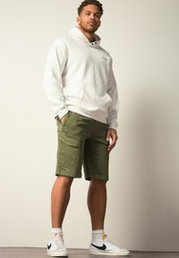 Sweat à capuche blanc avec une poche frontale, associé à un short cargo vert olive avec des poches latérales. L'ensemble se compose de sneakers blancs avec des accents noirs.