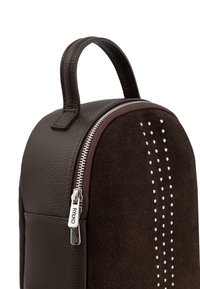 Zaino in pelle marrone con finitura scamosciata testurizzata, dettaglio della cerniera argentata e borchie decorative lungo un lato. Forma compatta con manico superiore.