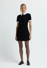 Donna con capelli biondi corti che indossa un mini abito nero con colletto e polsini bianchi, una cintura nera, collant trasparenti neri e scarpe nere con lacci.