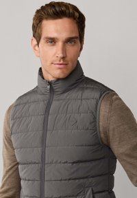 Grijze puffer bodywarmer met opstaande kraag, doorgestikt ontwerp, ritssluiting aan de voorkant en zijzakken, gedragen over een bruin shirt met lange mouwen.