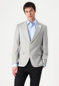 Lichtgrijze op maat gemaakte blazer met notched revers, twee knopen en twee voorzakken. Gedragen over een blauwe overhemd met een witte accentbloem.