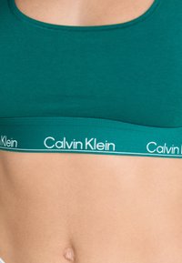 Teal sportsbh med en bred elastisk kant, der har hvid "Calvin Klein" branding. Glat materiale, scoop-hals og minimalistisk design.