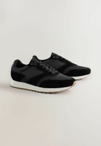Ein Paar schwarze Sneakers mit Wildleder- und Stoffeinsätzen, weißen Zwischensohlen und Außensohlen aus Gummi, auf einer neutralen Oberfläche platziert.