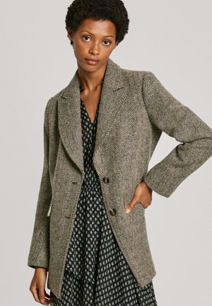 Manteau beige avec un motif en chevrons, à la coupe ajustée, avec des revers crantés et deux boutons, porté par-dessus une robe à motifs sombres. Texture lisse.