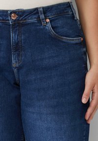 Ciemnoniebieskie dżinsy o dopasowanym kroju, z mosiężnymi guzikami, subtelnymi detalami przeszycia i klasycznym pięciopocketowym wzorem.