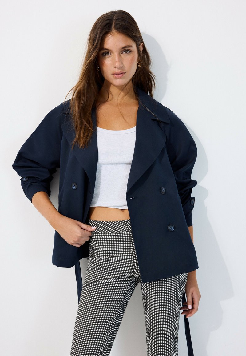 Young woman with long brown hair wearing a navy double-breasted jacket, white crop top, and black and white checkered pants.